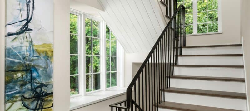 Modern staircase with black railings in a bright, airy room featuring large windows, an abstract painting, and white walls, creating an elegant and open space